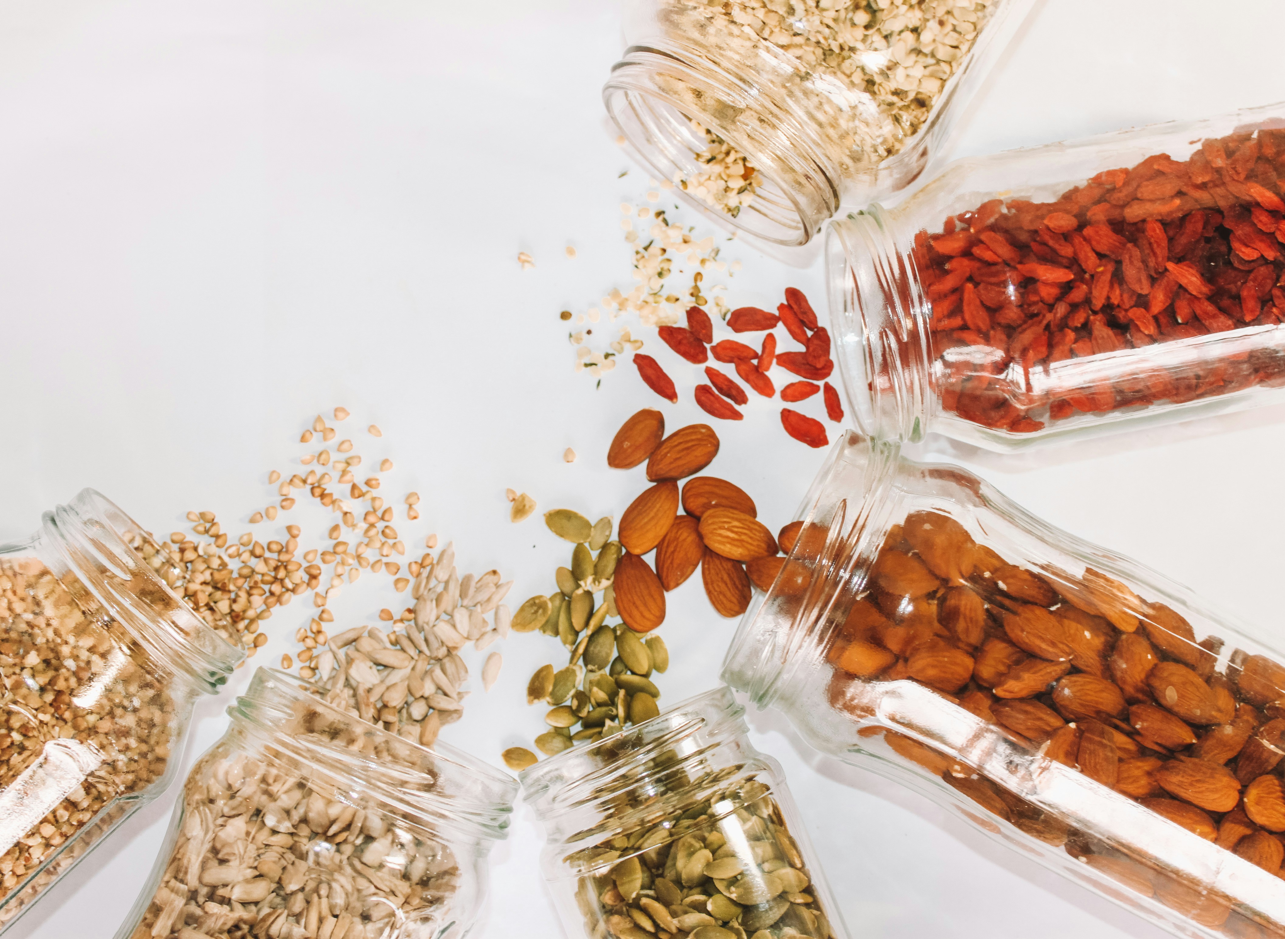 Assorted nuts and seeds spilling from glass jars onto a white surface, arranged in a circular pattern. The scene is bright and clean, evoking a sense of freshness and abundance. No visible text.